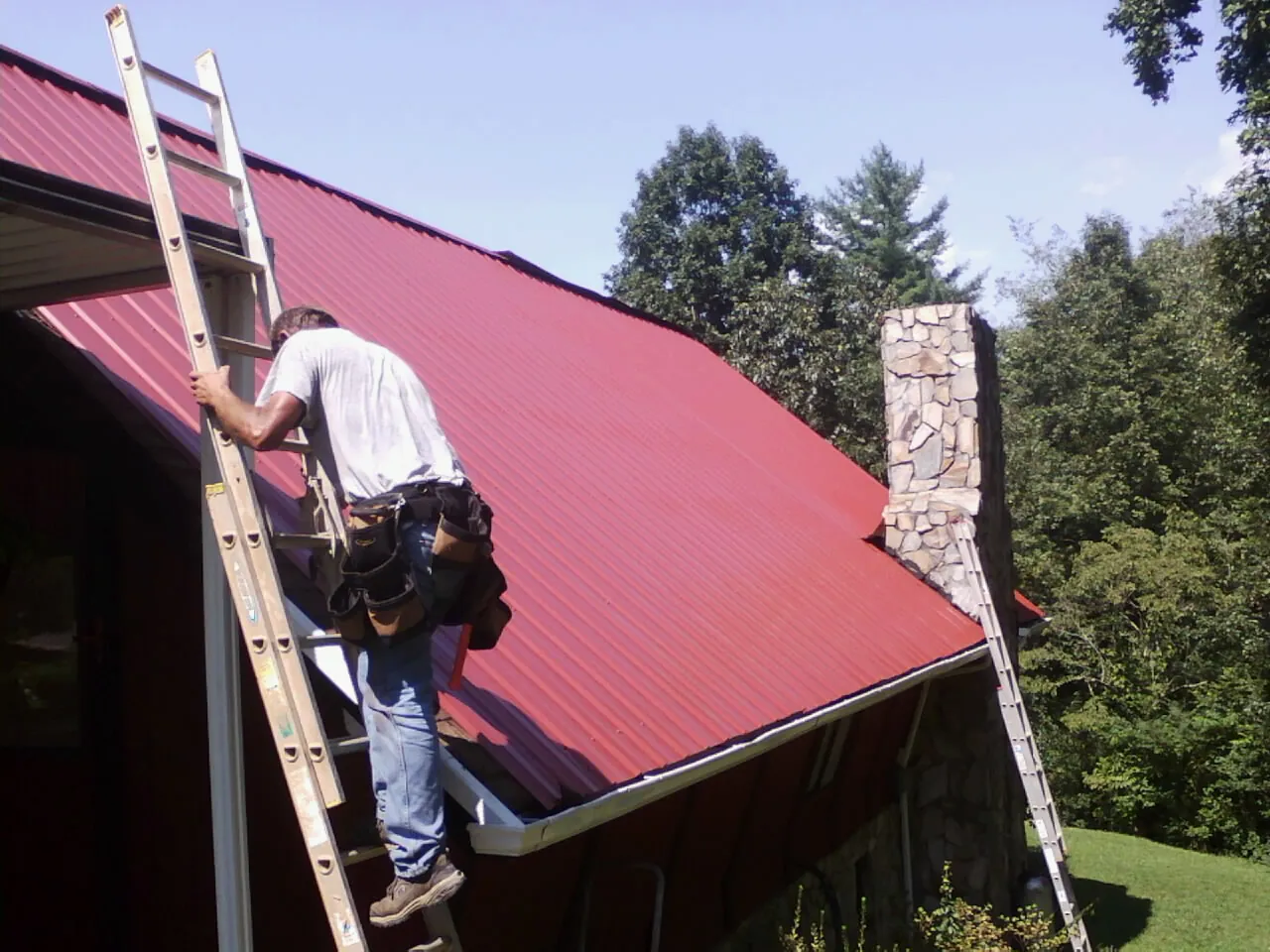 Roofer on a ladder beside a red metal roof and stone chimney — Soffit & Fascia Repair by Roofing Brothers in Alton